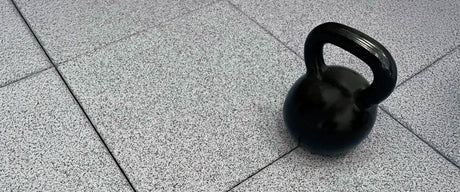 A black cast iron kettlebell sitting on gray rubber floor tiles in a gym.