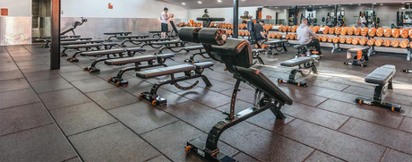 Free weights area of a gym with benches, dumbbells, and rectangular rubber floor tiles.