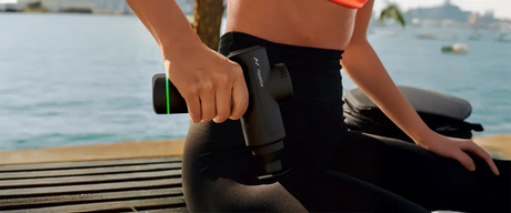 A woman using a Hyperice Hypervolt massage gun on her right upper thigh/hip area.