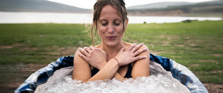 A woman sitting in an icebath with her eyes closed, in a green pasture by a lake.