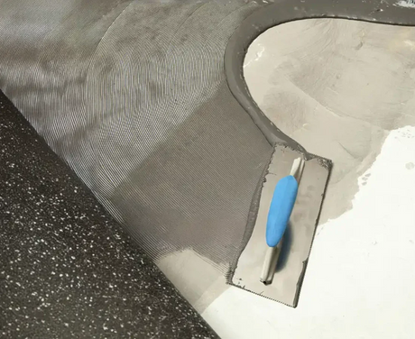 A trowel spreading adhesive on a cement floor with a rubber piece of flooring stuck down.