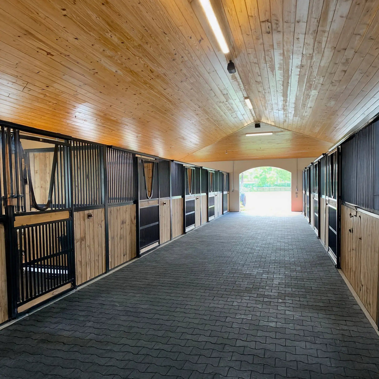 A clean, well-lit horse stable with wooden walls and ceiling, black metal stall doors, and an ECORE Athletic ECORE Rubber Paver 1" (24 mm) center aisle. Sunlight streams in from an open doorway at the far end.