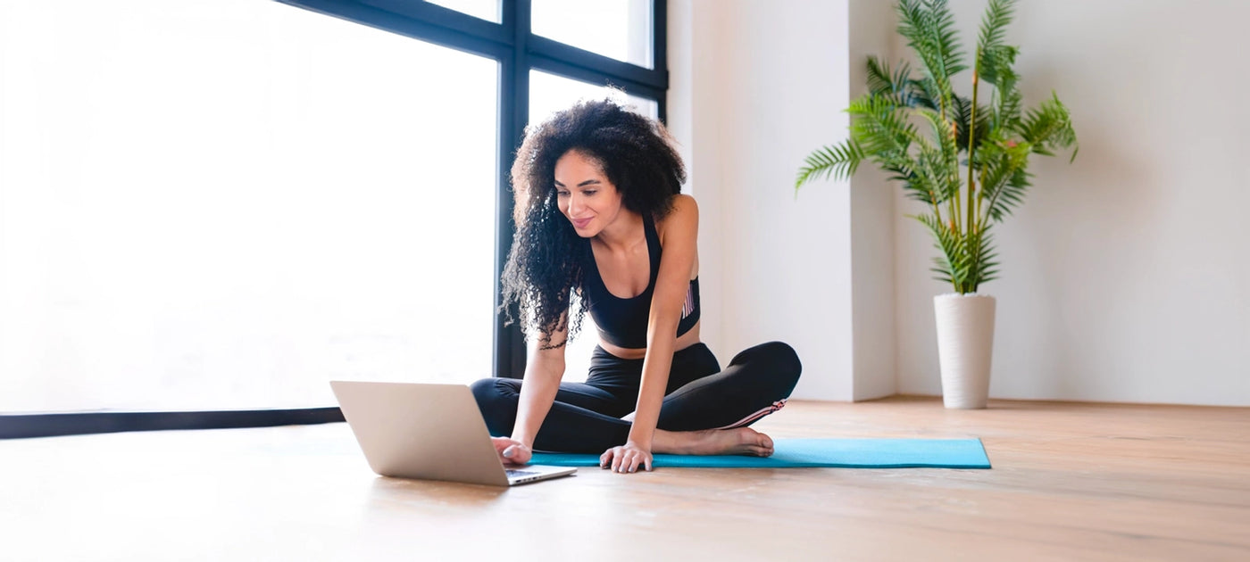 Woman sitting on a blue yoga mat on a hardwood floor while looking at her laptop which is on the floor in front of her.