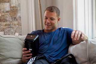 Christian McCaffrey sitting on a couch holding a control unit while wearing Normatec 3 Legs.