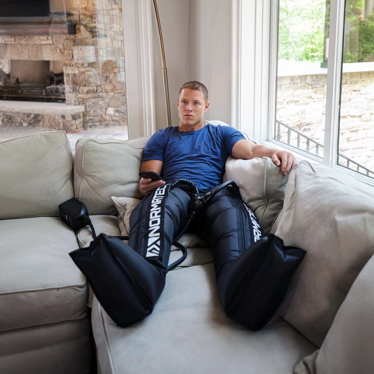 Christian McCaffrey wearing a pair of Normatec Leg inflatable leggings while using his phone and laying on the couch.