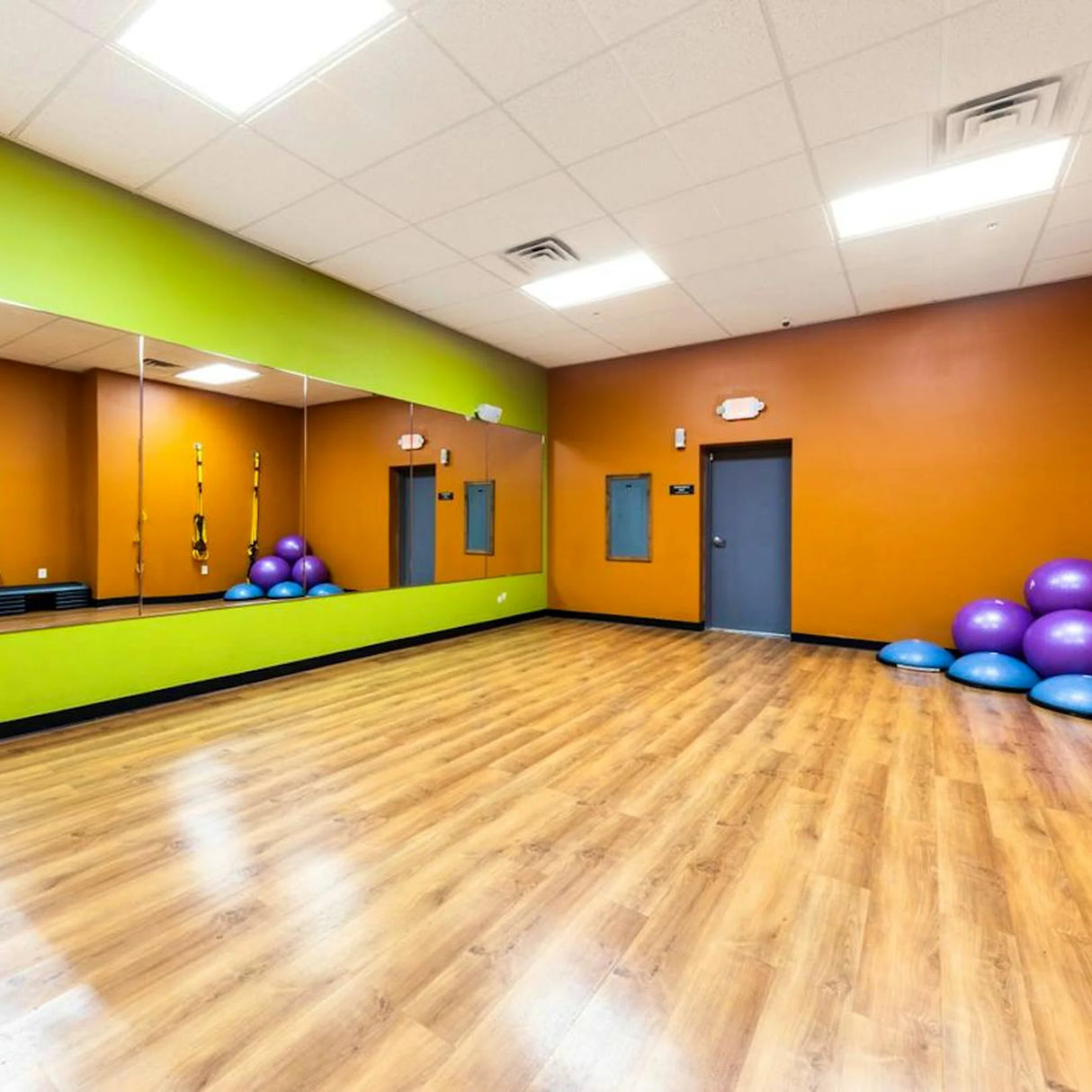 A spacious fitness studio with wood flooring cleaned by ECORE E-Cleaner All Purpose Floor Cleaner from ECORE Athletic; orange and green walls, large mirrors, purple exercise balls, blue balance discs in the corners, and bright ceiling lights.