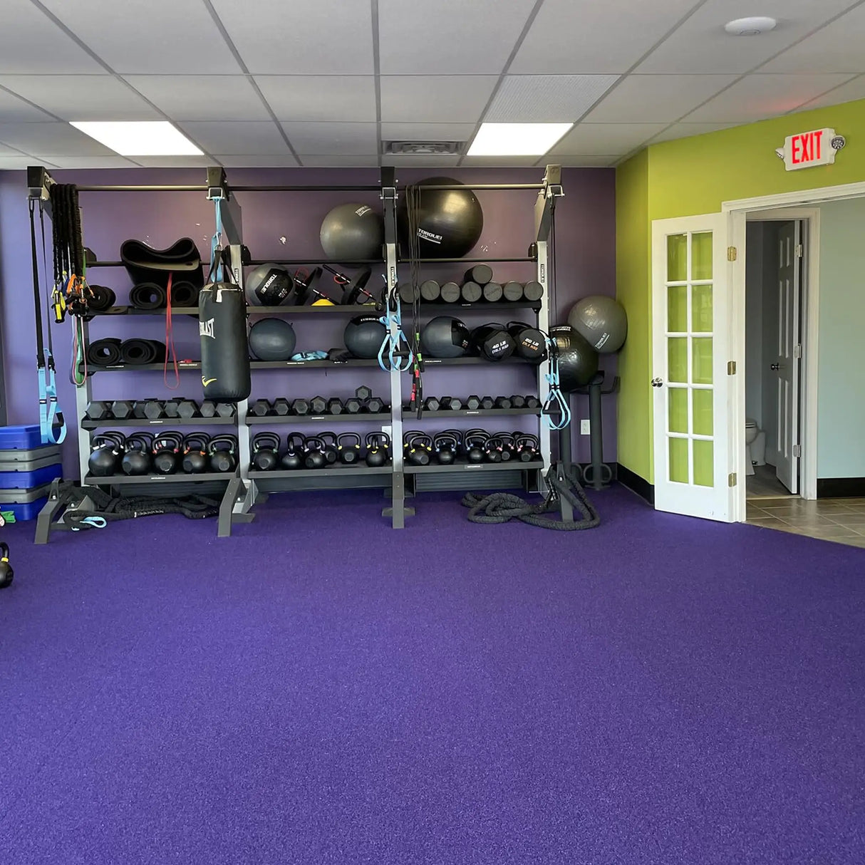 A gym with purple carpet and colorful walls features racks for kettlebells, weights, yoga mats, fitness gear, and a punching bag. Clean floors easily with ECORE Athletic's ECORE E-Cleaner All Purpose Floor Cleaner.