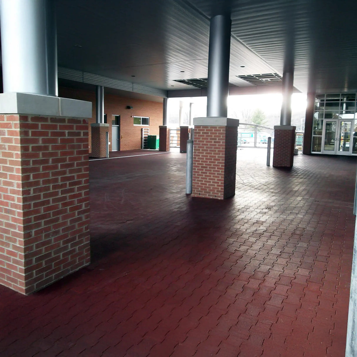 Covered walkway with large brick columns and ECORE Athletic's 1" ECORE Rubber Paver flooring outside a modern building with glass doors and windows. The area is empty and well-lit by natural light.