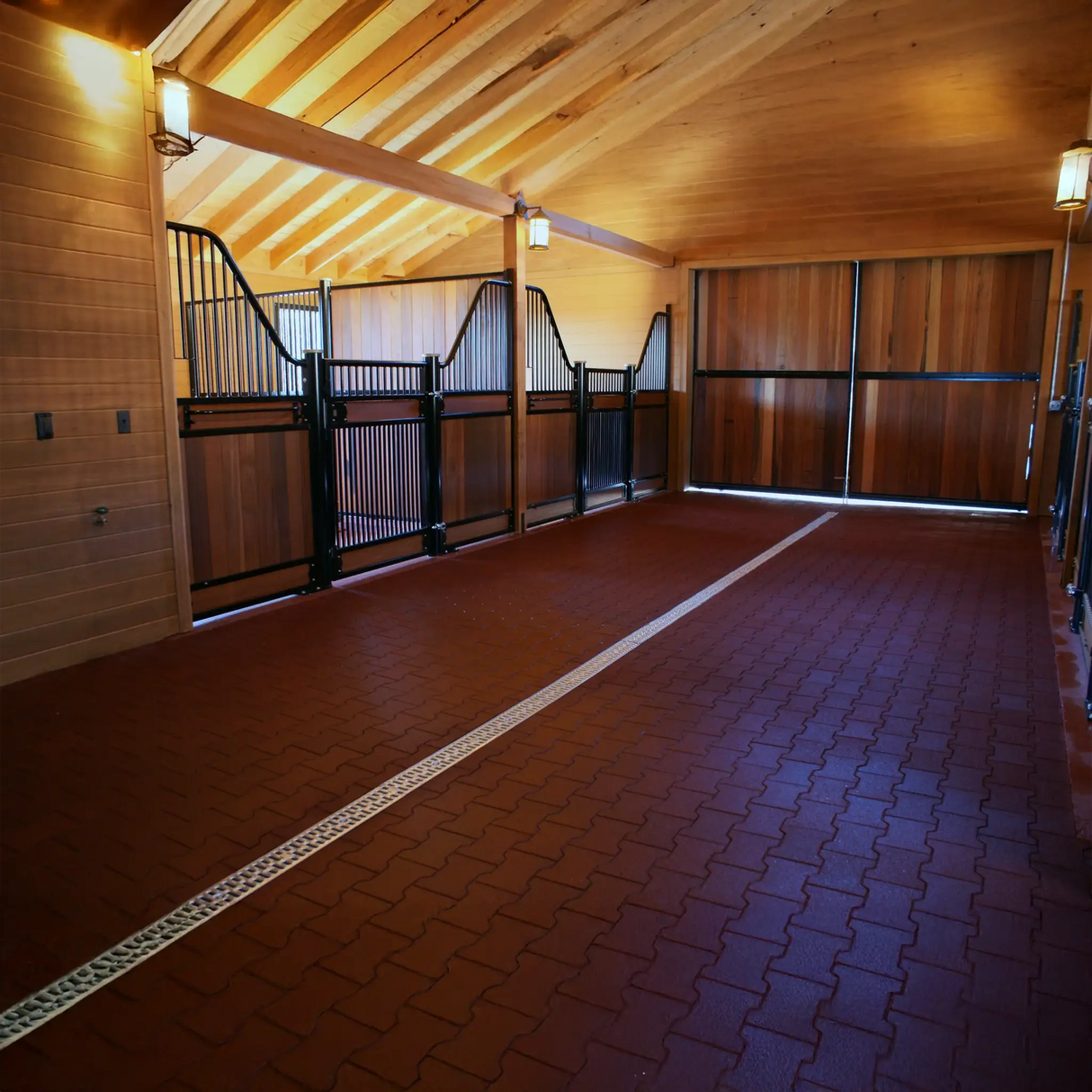 A clean, spacious horse stable with wooden walls and exposed beams features ECORE Athletic ECORE Rubber Paver, 1" (24 mm) flooring. Three empty stalls with black metal gates line the left side, leading to large wooden doors at the end.