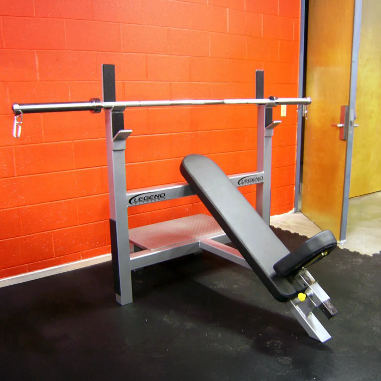 Gym bench and barbell rack against an orange brick wall.