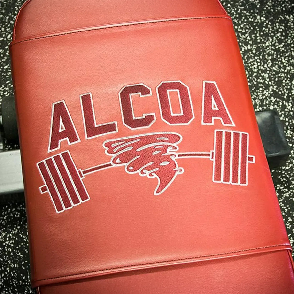 Red weightlifting bench with 'ALCOA' logo featuring a tornado and barbell embroidered on it on a black with white speckled gym floor background.