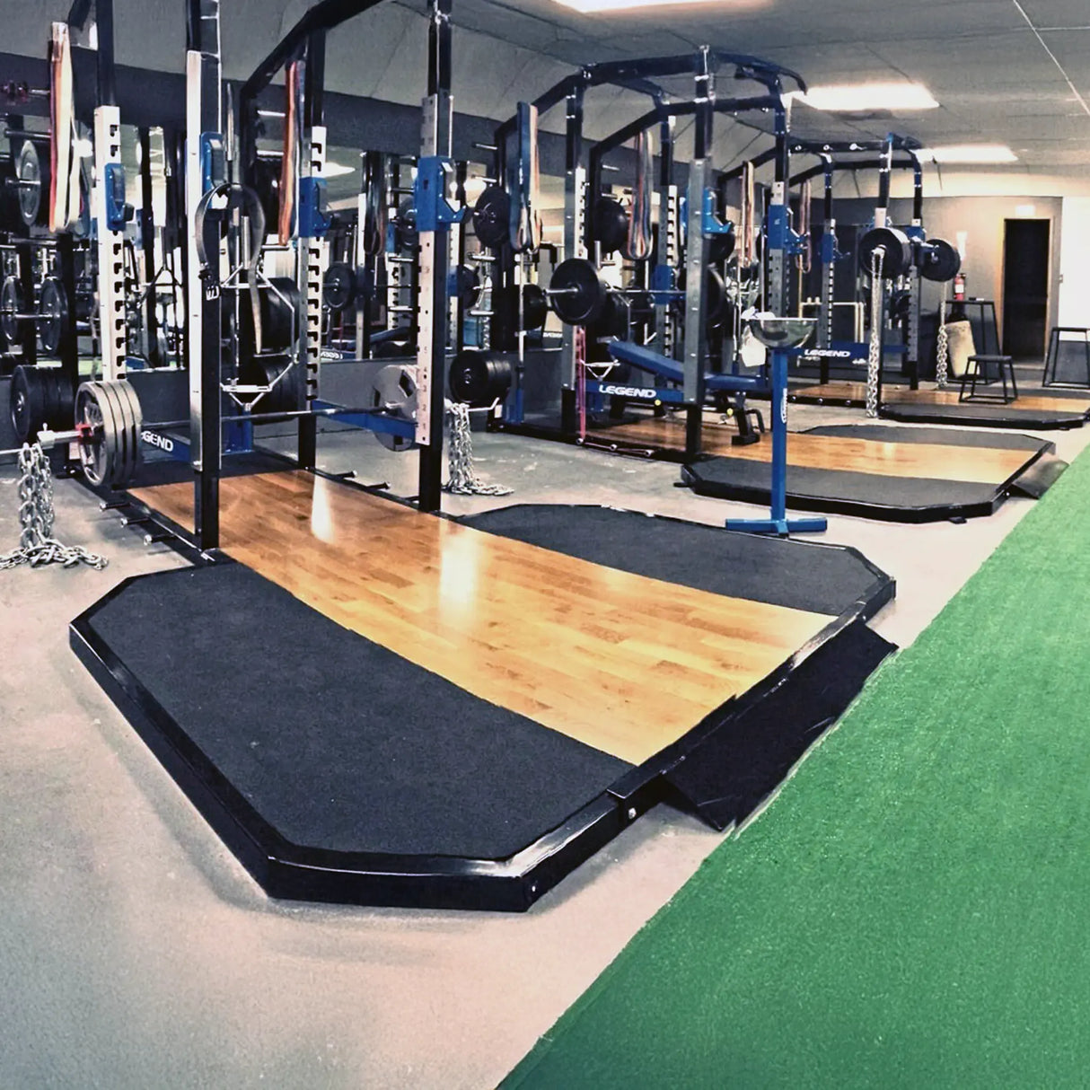 Gym interior with squat racks connected to weightlifting platforms next to a large piece of artificial turf