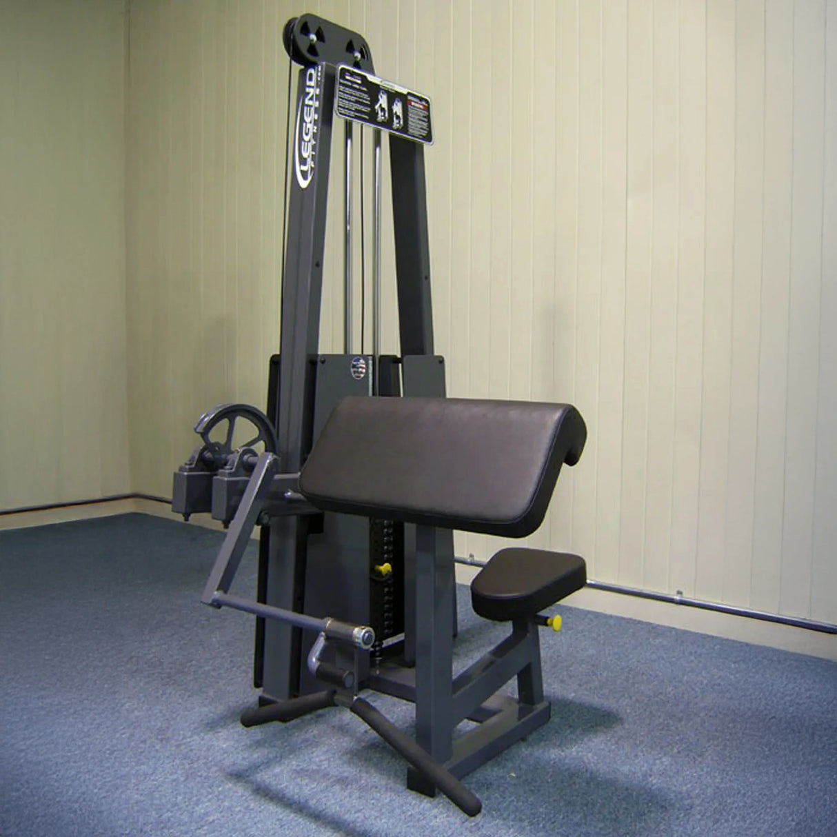 The Legend Fitness 908 Arm Curl Machine by Legend Fitness, in black and gray with padded seat and armrest for preacher curls, sits against a light-paneled wall on blue carpet in an empty room.