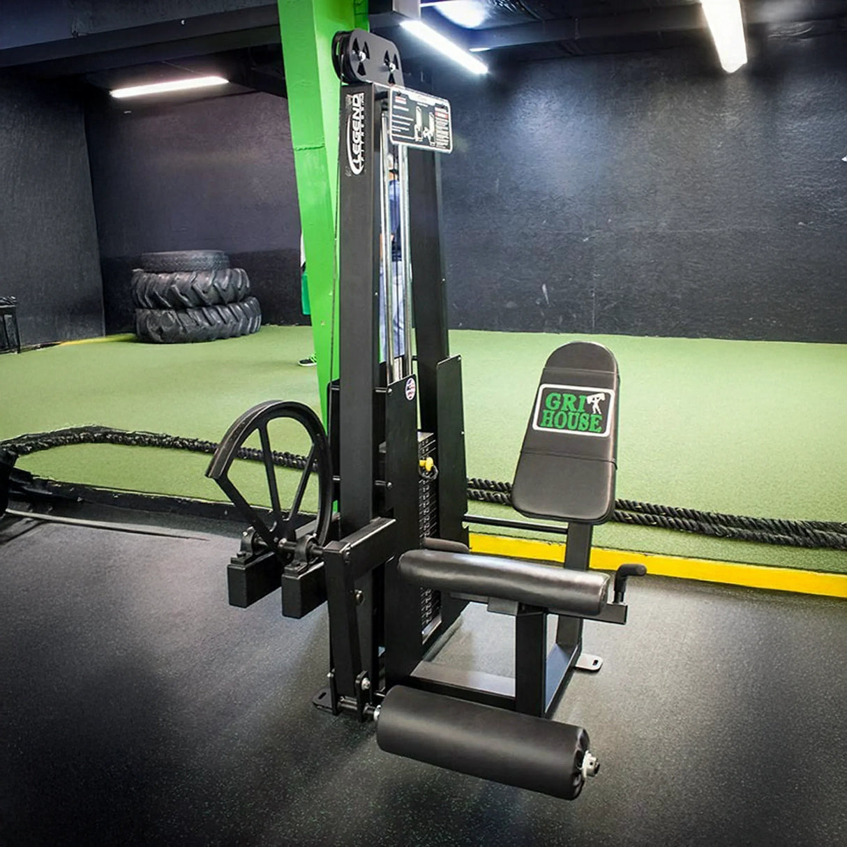 A Legend Fitness 911 Leg Extension Machine with a padded seat and leg rollers is in the gym, branded on the backrest. The workout area features black flooring, green turf, and stacked tires in the background.