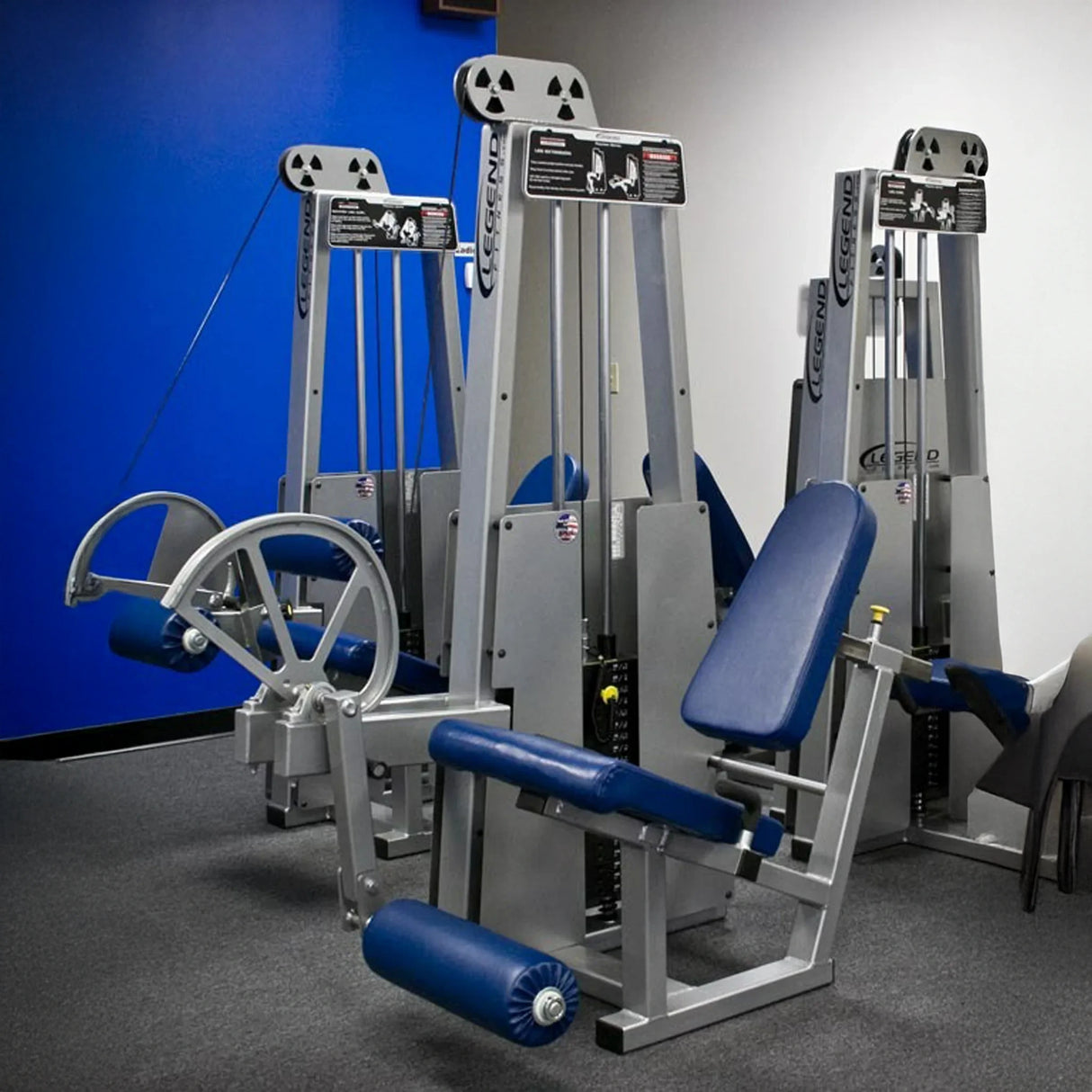 Two Legend Fitness 911 Leg Extension Machines by Legend Fitness, in blue and silver with padded seats and weights, are set up in a gym room with blue and white walls, gray carpet, and chairs partly visible on the right.