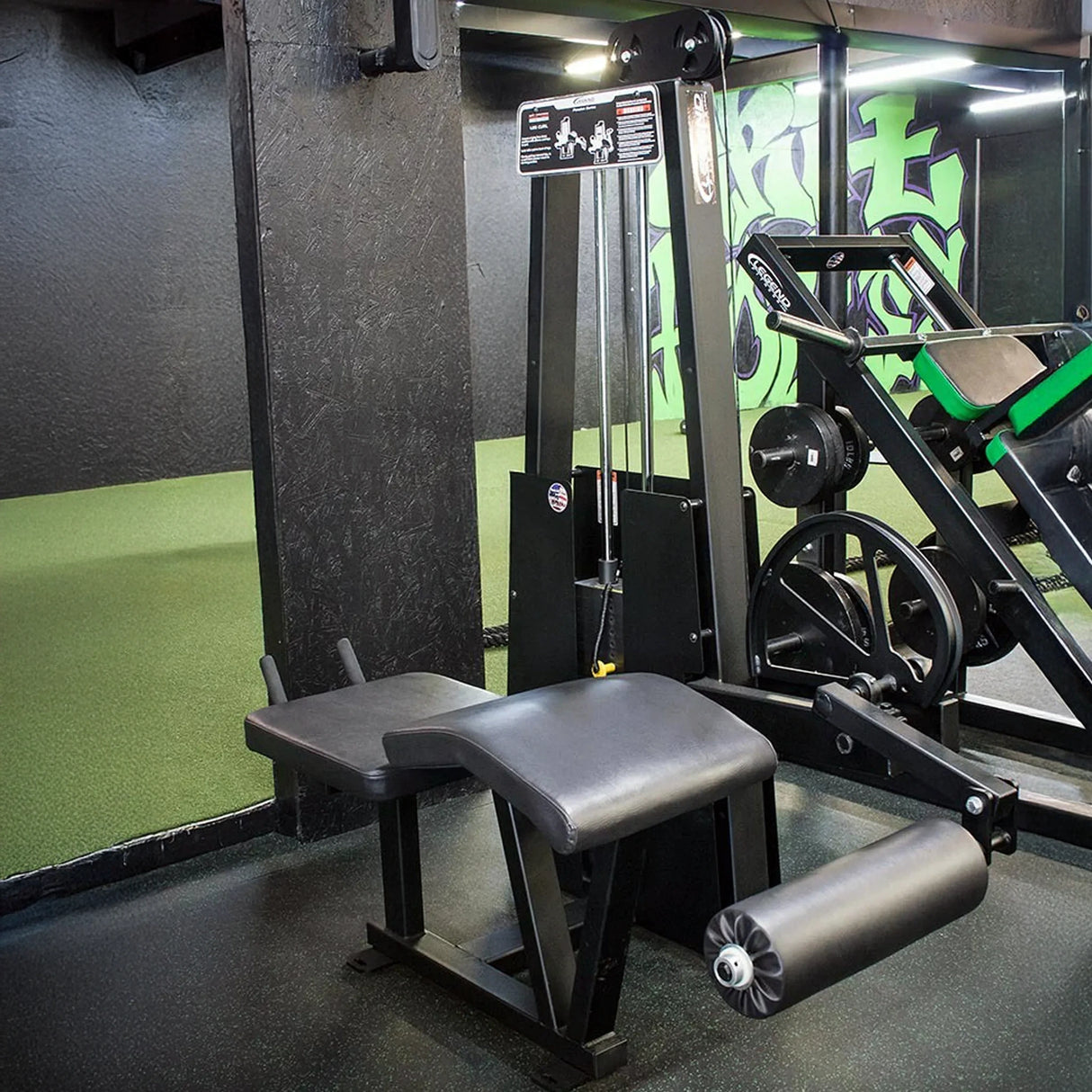 A Legend Fitness 912 Leg Curl Machine by Legend Fitness is set up in a gym featuring green and black walls, mirrored surfaces, and other fitness equipment in the background.