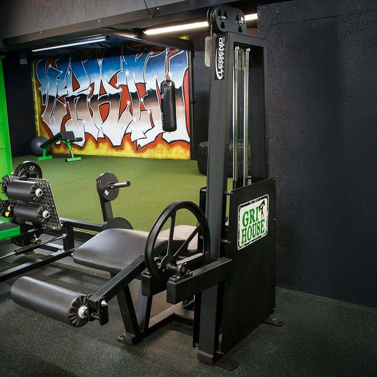 A Legend Fitness 912 Leg Curl Machine with a black seat is in a gym featuring green turf flooring, a graffiti mural on the wall, and a Grit House sign on the machine's side.