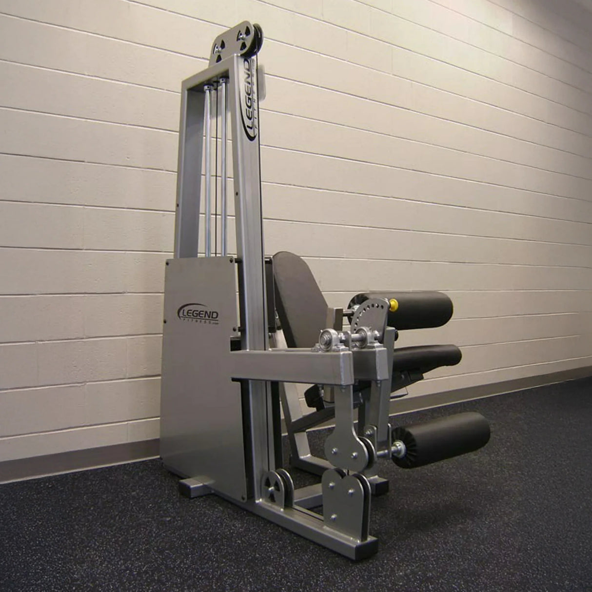 The Legend Fitness 918 Leg Extension / Leg Curl Combo Machine by Legend Fitness, featuring padded black rollers and a silver frame, is set against a beige brick wall on a dark speckled gym floor.