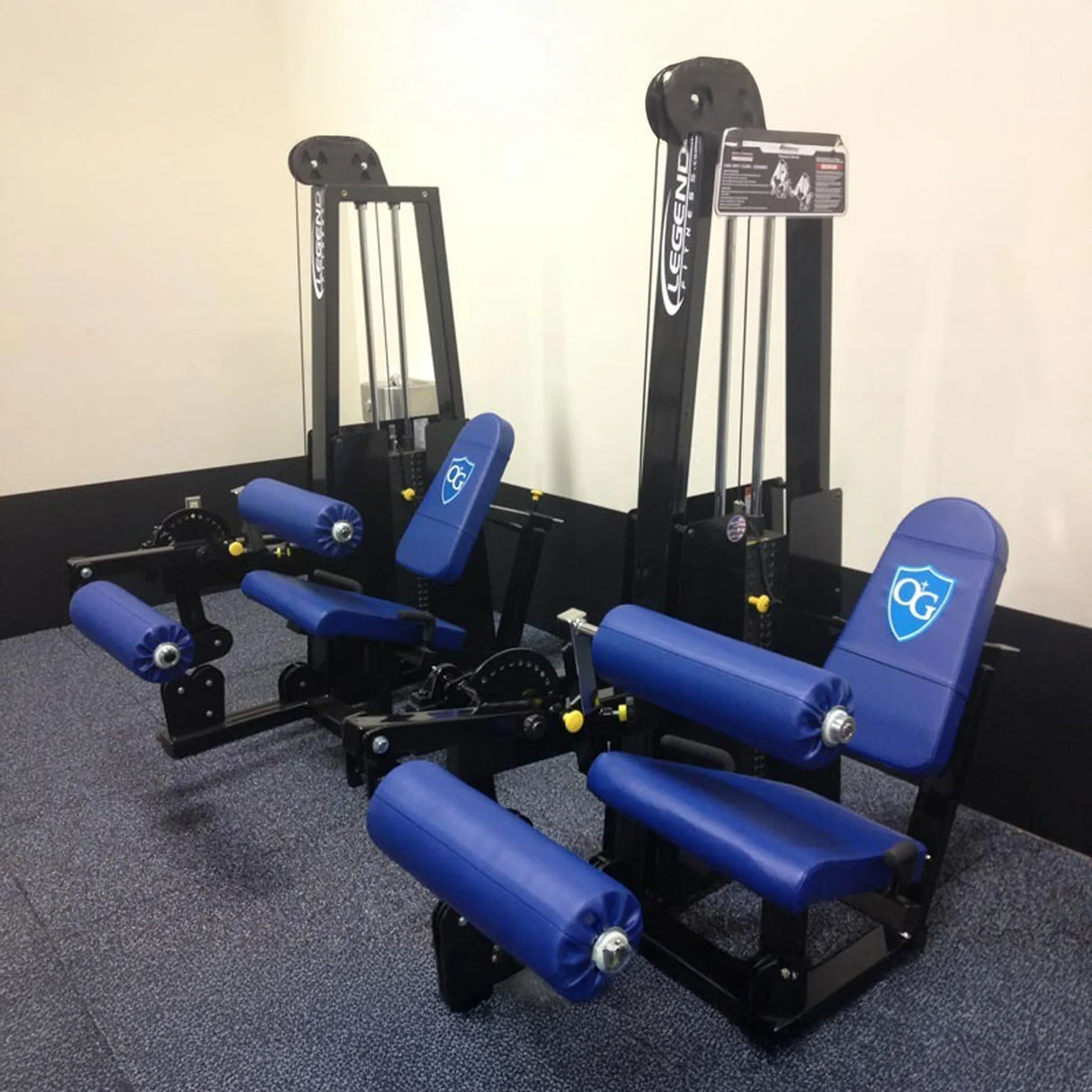 Two Legend Fitness 918 Leg Extension/Leg Curl Combo Machines in blue and black, featuring padded seats and rollers, are placed side by side on grey carpet against white walls.