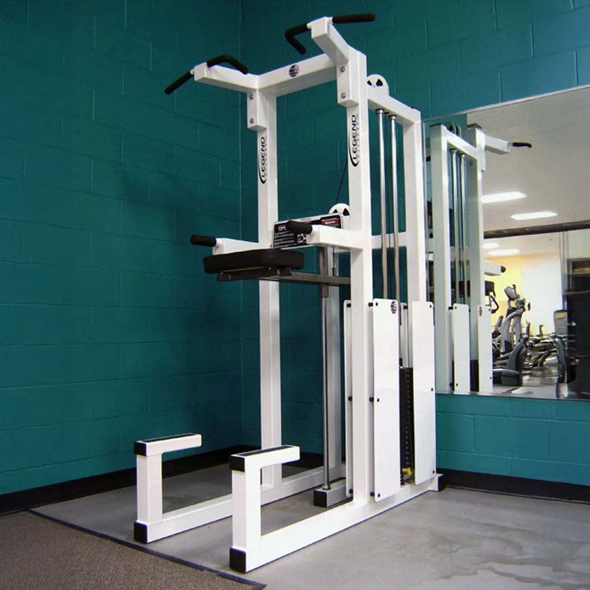 A Legend Fitness 933 Dip / Chin-Up Assist Combo Machine by Legend Fitness stands in the corner of a gym with teal walls and a large mirror, reflecting other fitness equipment in the background.