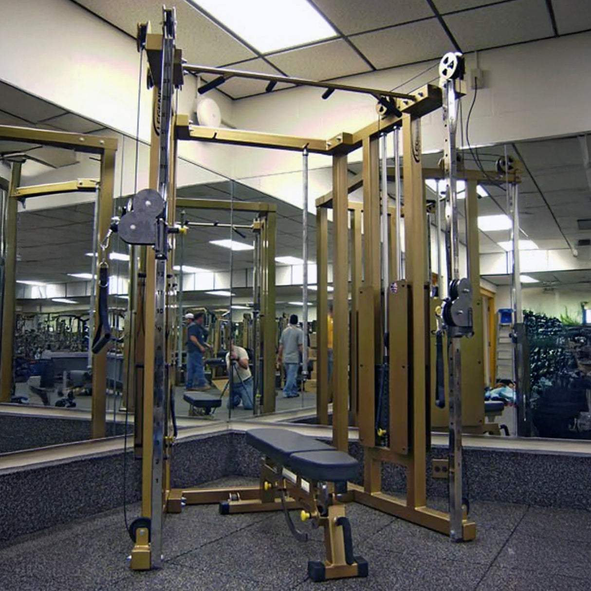A Legend Fitness 953 Functional Trainer Machine in gold and black, with an adjustable bench, stands before gym mirrors, reflecting the equipment and active members in the background.