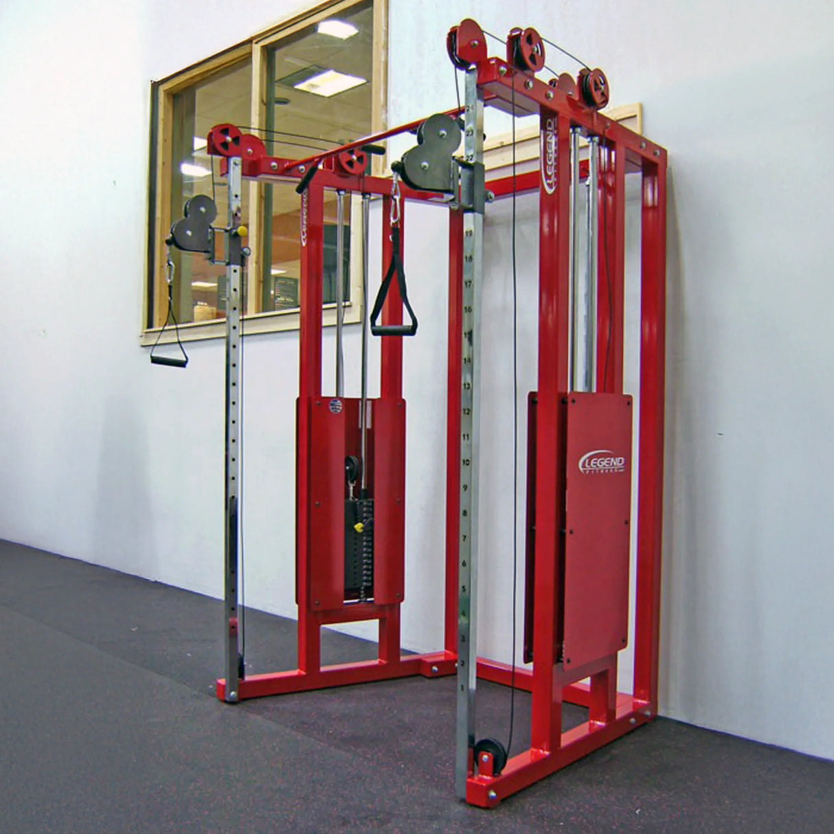 A Legend Fitness 953 Functional Trainer Machine with adjustable pulleys stands against a white gym wall near a large interior window. The floor beneath the machine features dark rubber mats.