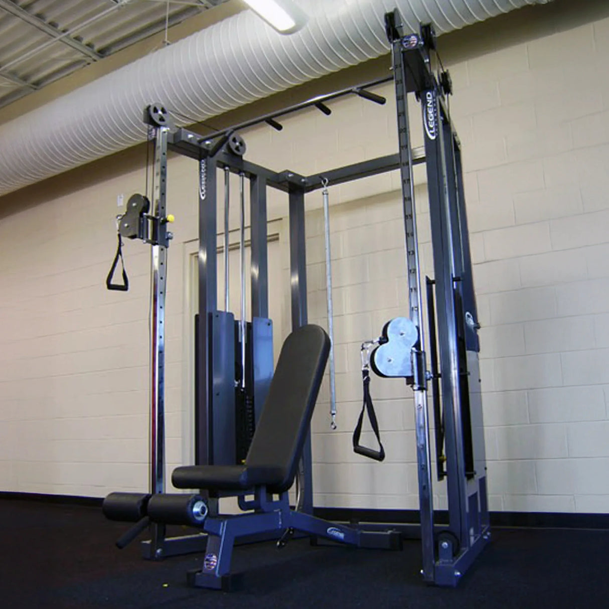 The Legend Fitness 953 Functional Trainer Machine by Legend Fitness, featuring a bench, adjustable pulleys, weights, and handles, sits in a spacious fitness room with beige walls and exposed ceiling ducts.