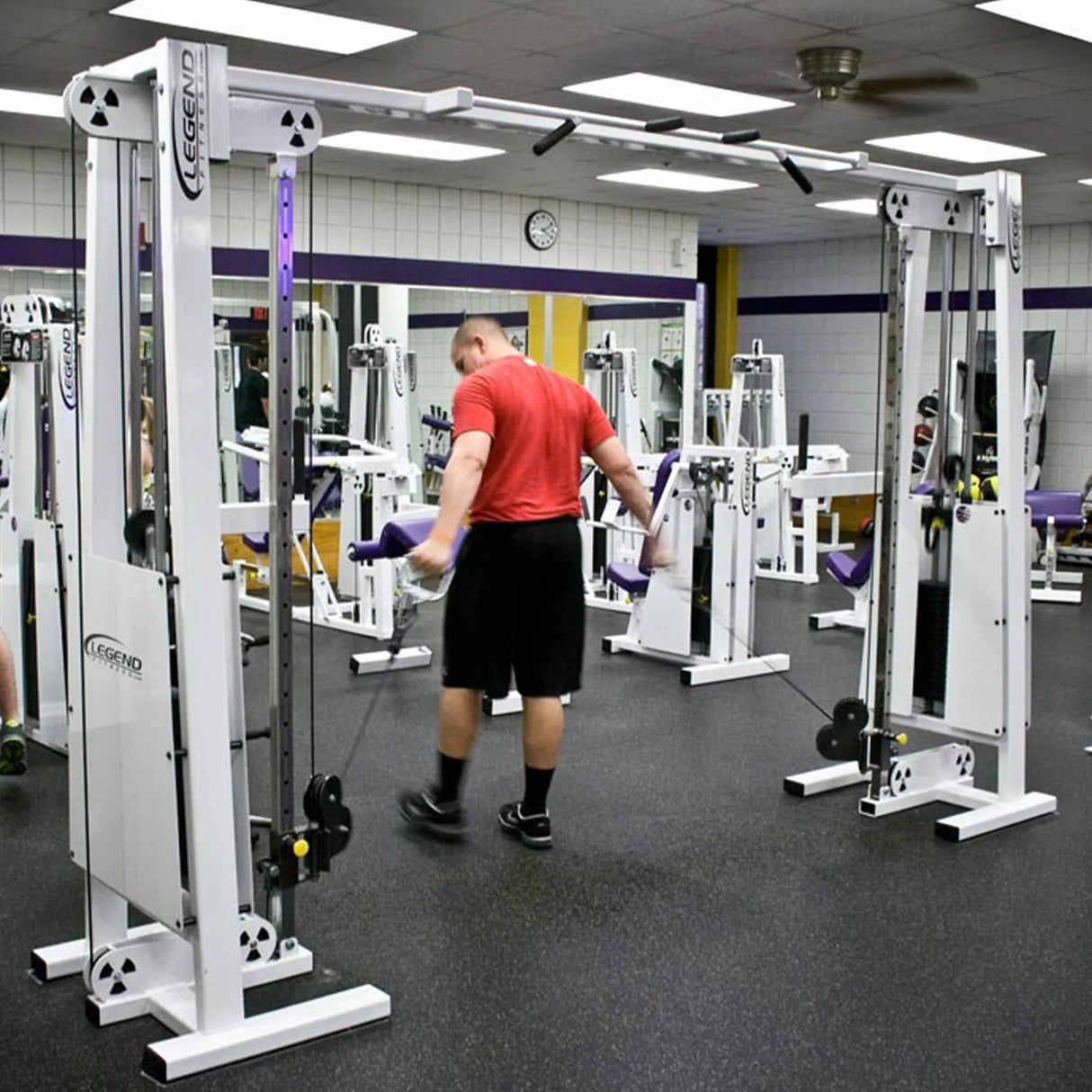 A man in a red shirt and black shorts uses the Legend Fitness 954 Cable Crossover with Adjustable Pulleys at a gym equipped with mirrors, a wall clock, and various exercise machines.
