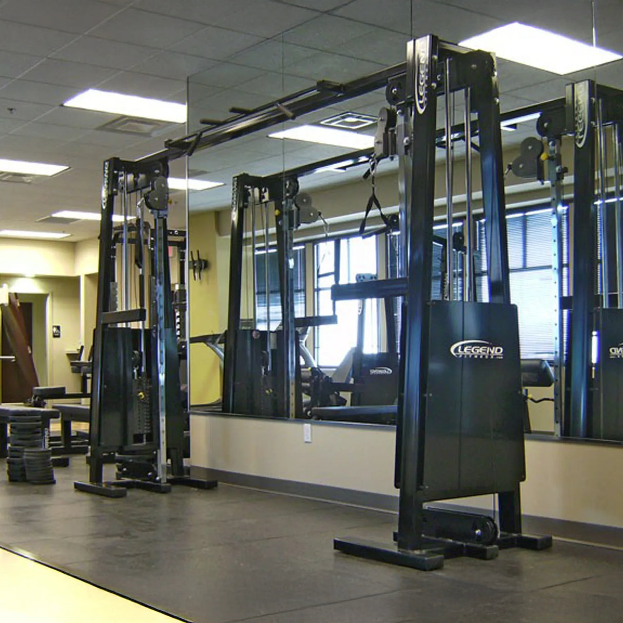 A modern gym features the Legend Fitness 954 Cable Crossover with Adjustable Pulleys in front of a large mirror. The room has rubber flooring, neutral walls, natural light from windows, and other gym equipment and weights visible in the background.