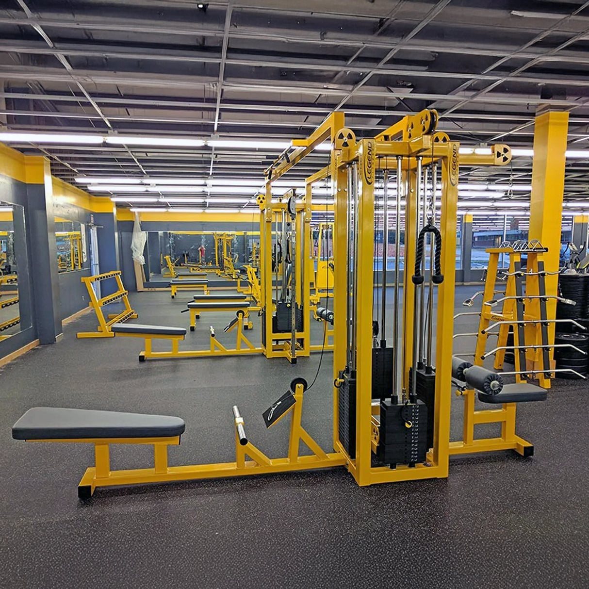 A spacious, well-lit gym featuring the Legend Fitness 959 Eight-Stack Jungle Gym in yellow and black, with benches, weight machines, and wall mirrors; the area is empty under bright overhead fluorescent lights.