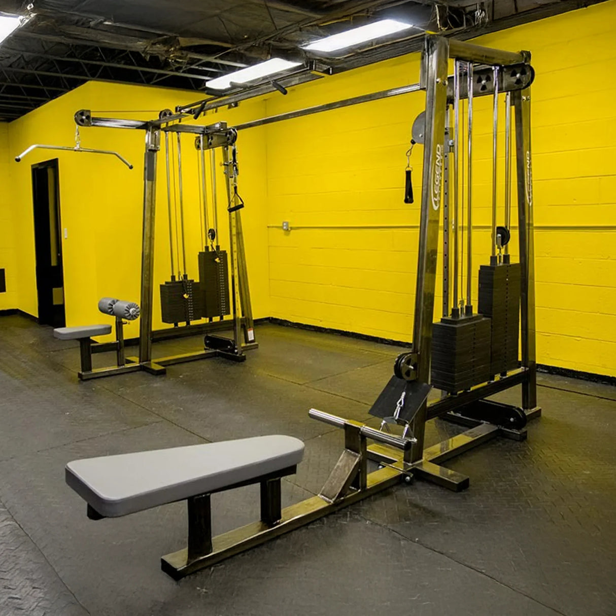 A clean and organized gym with yellow walls, black flooring, padded benches, and two Legend Fitness 960 Cable Crossover Plus Machines featuring cables, pulleys, and weight stacks.