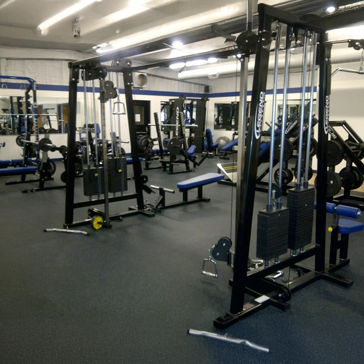 A clean, well-lit gym features the Legend Fitness 960 Cable Crossover Plus Machine among weight machines, benches, barbells, and free weights. Large mirrors line the walls and black rubber mats cover the floor.