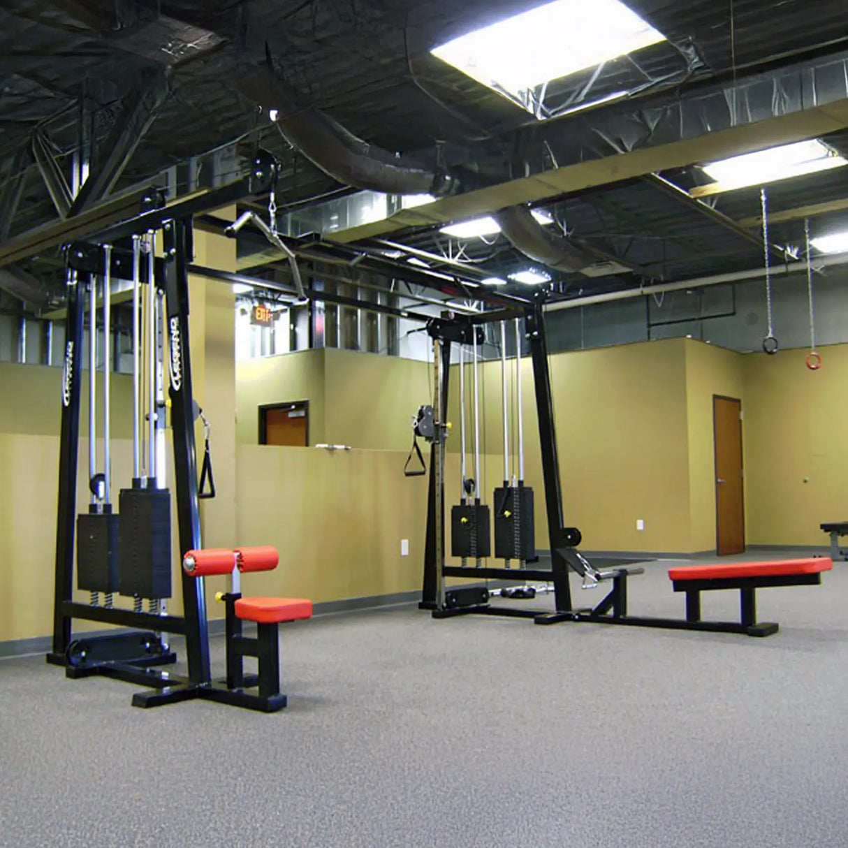 A spacious gym room features two Legend Fitness 960 Cable Crossover Plus Machines, each equipped with pulleys, weights, and red-padded benches on a carpeted floor, set against tan walls and exposed ceiling ducts.