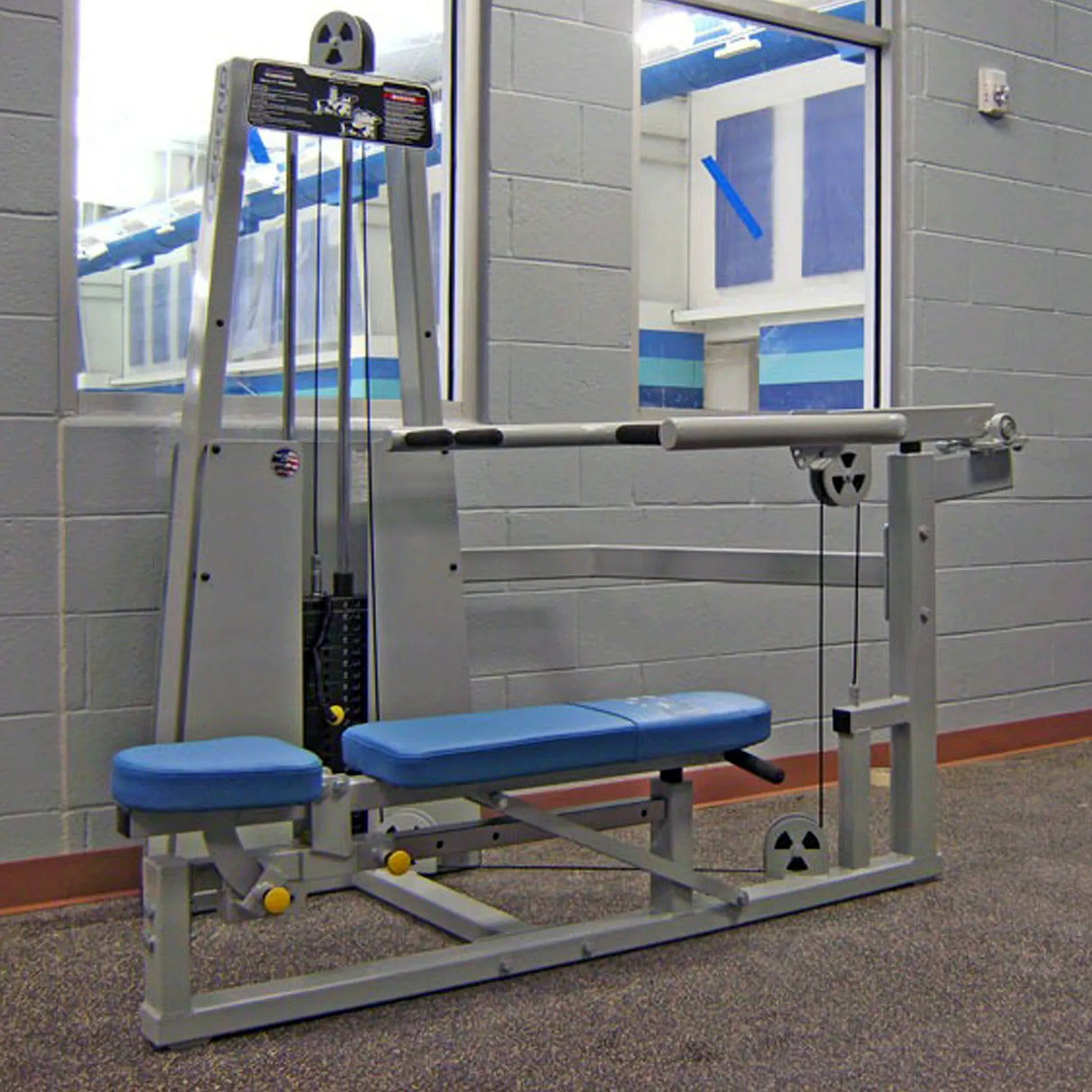 The Legend Fitness 963 Multi-Press Combo Machine by Legend Fitness, featuring blue padded seats and a silver frame, is set in a gym with gray walls and a large mirror. It uses cables and pulleys for resistance exercises.