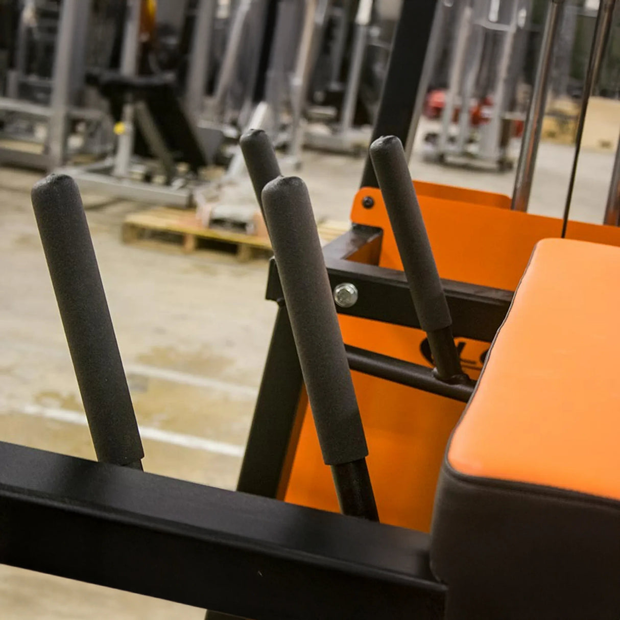 Close-up of foam-padded handles on the orange and black Legend Fitness 974 Pro Reverse Back Extension Machine, with other gym equipment blurred in the background.