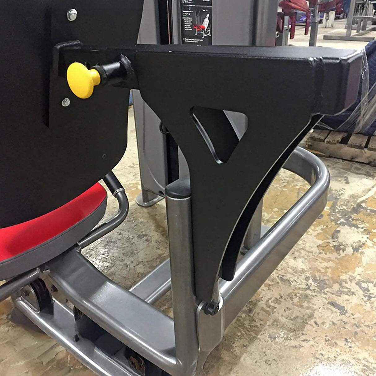 Close-up of a gym machine with a red seat and black mounting arm on a concrete floor.