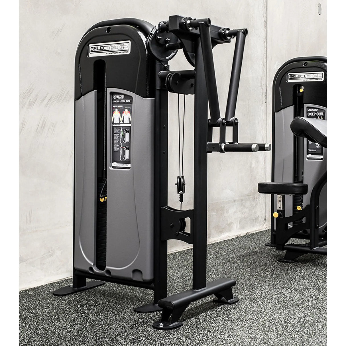 The Legend Fitness SelectEDGE 1113 Standing Lateral Raise Machine by Legend Fitness, in black and gray with illustrated instructions, is set on a speckled gym floor against a plain light-colored wall for upper body workouts.