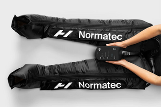 An overhead view of someone sitting with their legs out straight wearing an inflated pair of Normatec 3 Legs while holding the control unit in their hands.