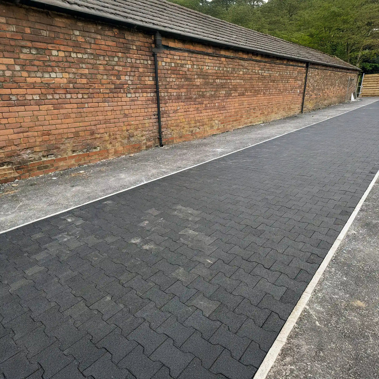 A pathway paved with ECORE Rubber Paver, 1" (24 mm) by ECORE Athletic runs alongside a long red brick wall with light concrete edging; greenery and trees can be seen in the background.
