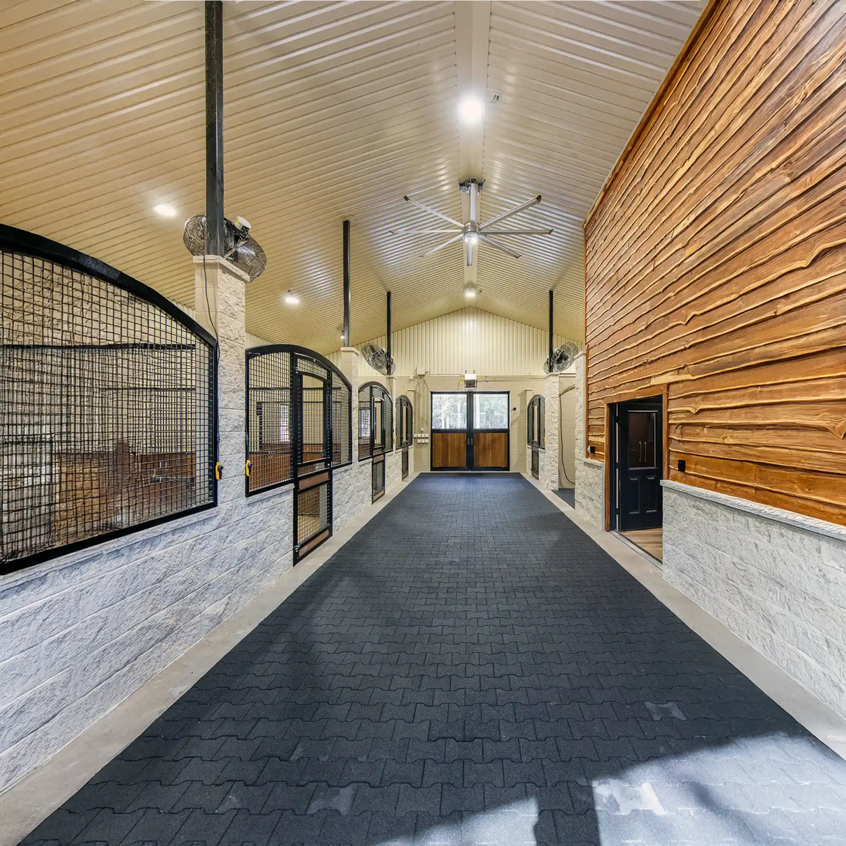 This wide, clean horse stable features ECORE Athletic’s 1" (24 mm) ECORE Rubber Paver flooring, white walls, black stall doors, wood panels on the right, a high ceiling with fans, and large double doors welcoming natural light.