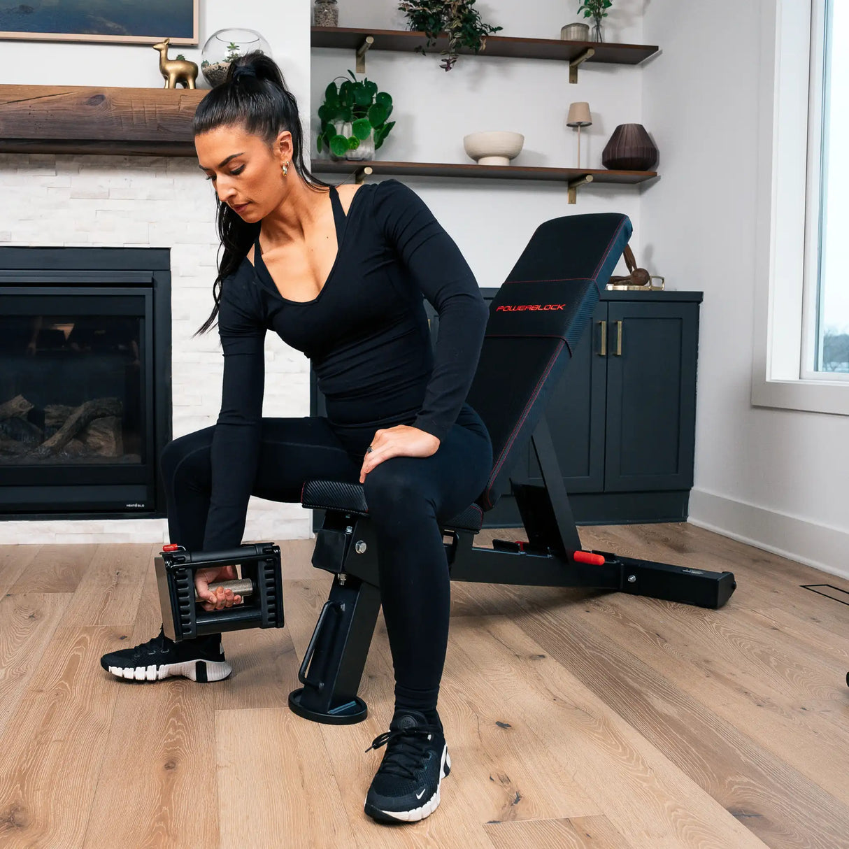 A woman curling a PowerBlock dumbbell while sitting on a black PowerBlock utility bench in the upright position