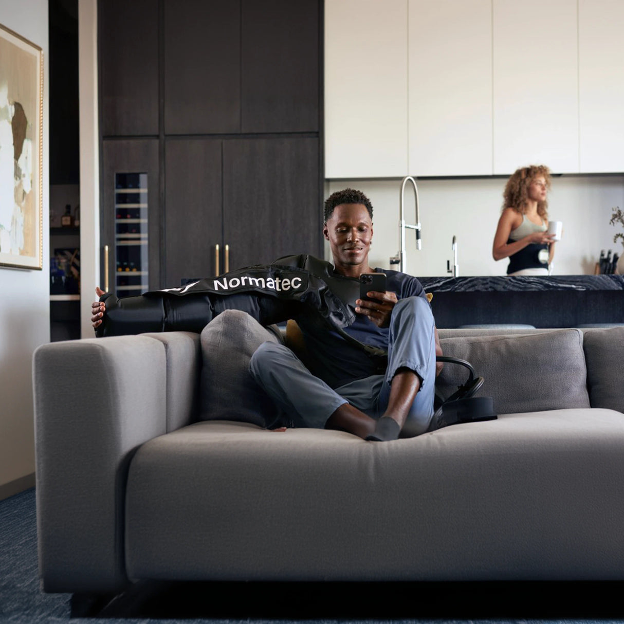 A man wearing a Normatec arm compression sleeve on his right arm while sitting on the couch and looking at his phone.