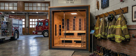 Large brown sauna with rich red and brown interior in a fire station with jackets on the wall, boots on the ground, and two fire trucks behind it. 
