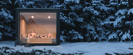 A lit up square sauna in the snow at night with a man and woman inside