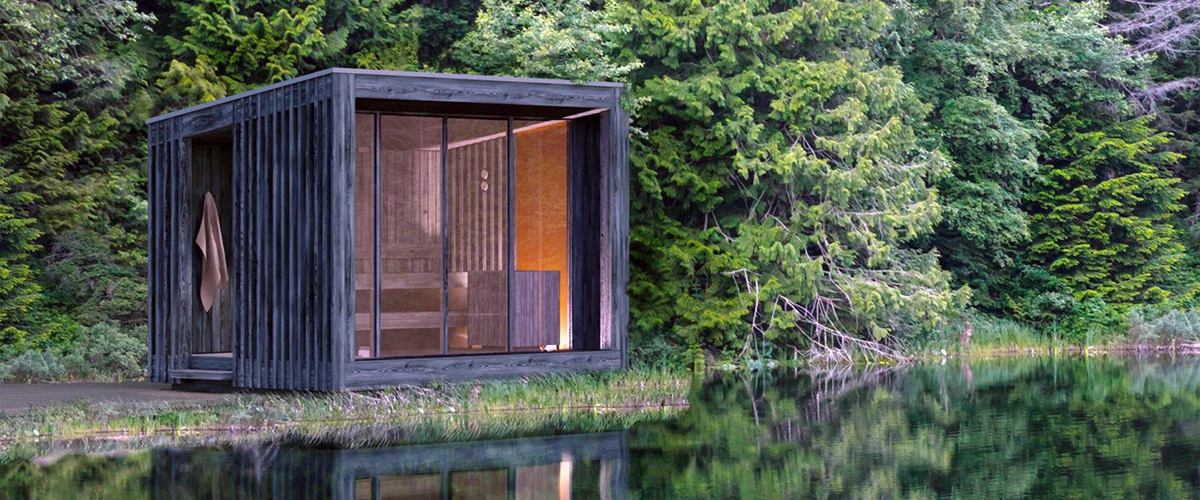 Black square sauna in front of some water with green pine trees behind it