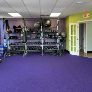 A gym with purple carpet and colorful walls features racks for kettlebells, weights, yoga mats, fitness gear, and a punching bag. Clean floors easily with ECORE Athletic's ECORE E-Cleaner All Purpose Floor Cleaner.