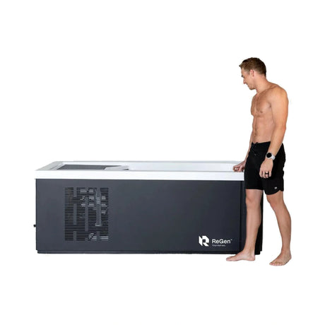 A rectangular cold plunge with a black exterior and white acrylic interior with an attractive brunette man in a pair of black swimming shorts walking along the side of it