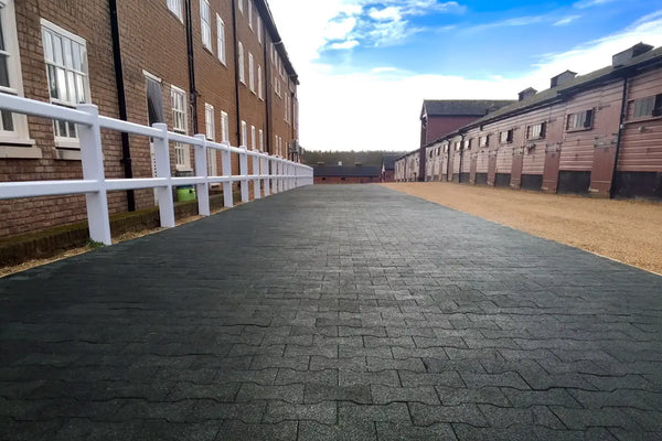 A long footpath made of black rubber pavers running between two long rows of buildings with a white wooden fence on the left between the footpath and the buildings
