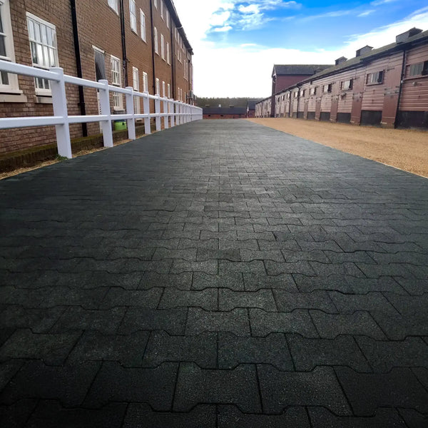A long footpath made of black rubber pavers running between two long rows of buildings with a white wooden fence on the left between the footpath and the buildings