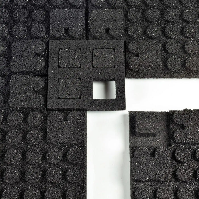 A close-up of ECORE Athletic’s ECORE Quad Blok Connector for 1" Tiles shows black foam tiles with textured surfaces in a grid. The central connector features four square cutouts, one missing to reveal the white background below.